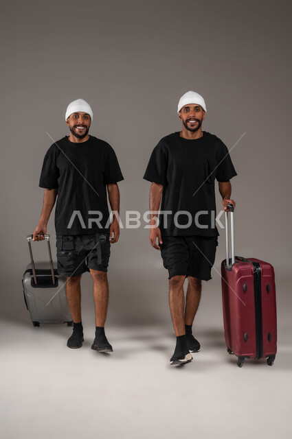 النظر باتجاه الكاميرا بسعادة ، الاستمتاع بقضاء أوقات ممتعة,  بورتريه لشابين توءم عربيين خليجيين سعوديين يرتديان الزي الكاجوال يمسكان بيديهما حقائب سفر، ايماءات الحماس والفرح،  صورة الجسم بالطول الكامل، خلفية رمادية