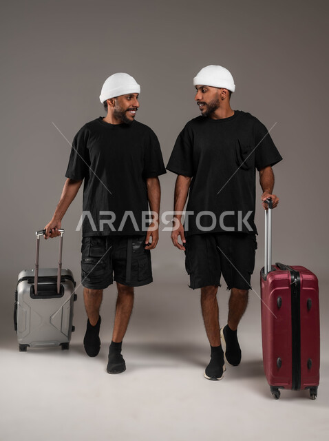 المحبة والود بين الاخوة، الاستمتاع بقضاء أوقات ممتعة,  بورتريه لشابين توءم عربيين خليجيين سعوديين يرتديان الزي الكاجوال يمسكان بيديهما حقائب سفر، ينظران إلى بعضهما بسعادة وسرور، صورة الجسم بالطول الكامل، خلفية رمادية