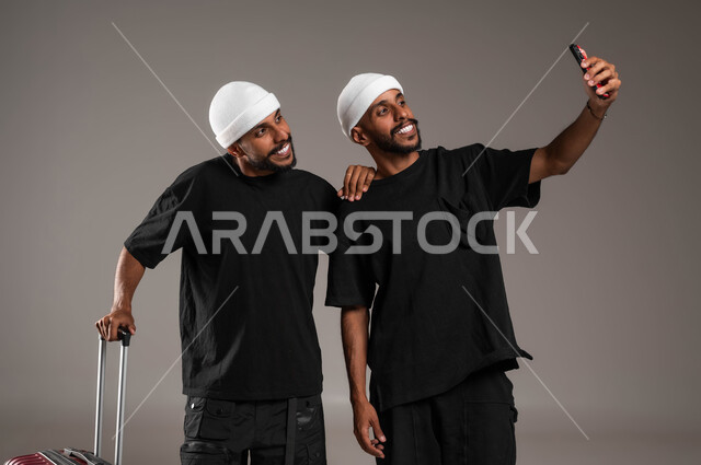 Using advanced technical devices, documenting happy moments, enjoying pleasant times, love and friendship between brothers, portrait of two young Saudi Gulf Arab twins wearing casual clothes, one of them holding a travel bag in his hand and the other taking a souvenir selfie, gestures of happiness and happiness, gray background.