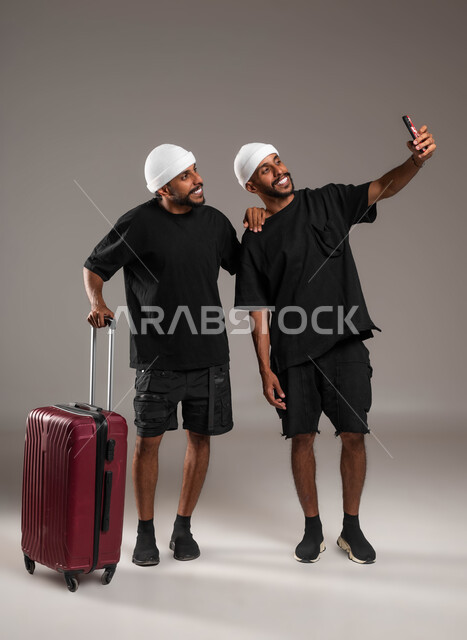 الاستمتاع بقضاء أوقات ممتعة, استخدام الأجهزة التقنية المتطورة، توثيق اللحظات السعيدة،  المحبة والود بين الاخوة، بورتريه لشابين توءم عربيين خليجيين سعوديين يرتديان الزي الكاجوال إحداهما يمسك بيده حقيبة سفر والآخر يقوم بالتقاط صورة السيلفي التذكارية، ايماءات السعادة والسرو، صورة الجسم بالطول الكامل، خلفية رمادية