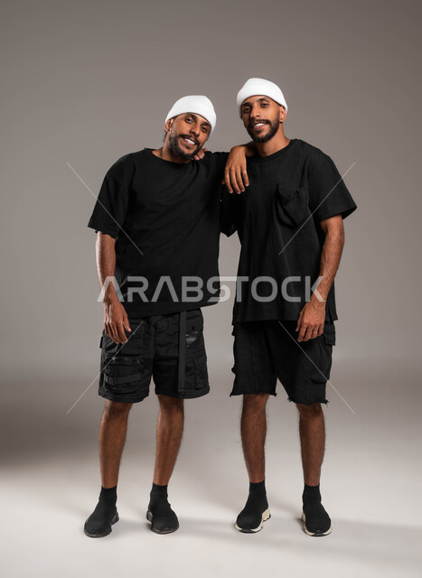 علاقات أخوية مترابطة، المحبة والود بين الاخوة، علاقات اجتماعية وصداقة ،ايماءات الفرح و السعادة، النظر باتجاه الكاميرا، الاستمتاع بقضاء أوقات ممتعة, ، بورتريه لشاب عربي خليجي سعودي يرتدي الزي الكاجوال يعانق أخيه التوءم ، صورة الجسم بالطول الكامل، خلفية رمادية