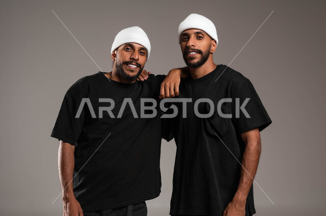 المحبة والود بين الاخوة، النظرباتجاه الكاميرا، علاقات أخوية مترابطة، ايماءات الفرح و السعادة،  الاستمتاع بقضاء أوقات ممتعة, بورتريه مقرب لشاب عربي خليجي سعودي يرتدي الزي الكاجوال يعانق أخيه التوءم ،خلفية رمادية