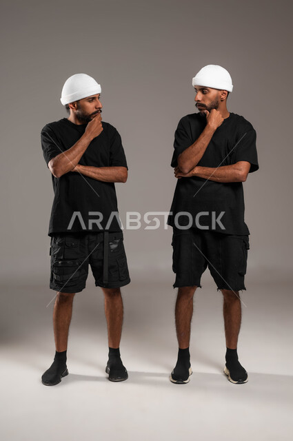 شرود الذهن والتفكير، انتظار شيء ما، محاولة إيجاد حل لمشكلة ما، الاحساس والشعور بالملل، بورتريه لاخين توءم عربييين خليجيين سعوديين مبتسمين يرتديان الزي الكاجوال وقبعة بيضاء ينظران لبعضهما بإيماءات التفكير، صورة لكامل الجسم، خلفية رمادية