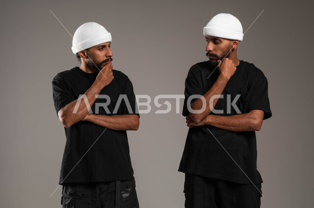 محاولة إيجاد حل لمشكلة ما، الاحساس والشعور بالملل، شرود الذهن والتفكير، انتظار شيء ما، ، بورتريه مقرب لاخين توءم عربييين خليجيين سعوديين مبتسمين يرتديان الزي الكاجوال وقبعة بيضاء ينظران لبعضهما بإيماءات التفكير، خلفية رمادية