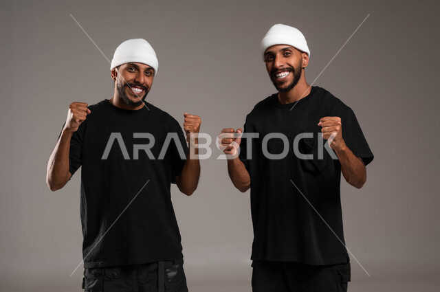 Feelings of love and friendliness between brothers, a feeling of joy and pleasure, a close-up portrait of two smiling young Saudi Gulf Arab twins wearing casual clothes and a white hat, raising hands high with gestures of victory, success and achieving goals, the concept of happiness in reaching goals, gray background