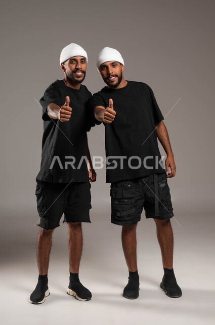 الاتفاق على أمر معين، النظر إلى الكاميرا بتعابير السرور، القبول والرضا بالنتيجة، ايماءات الاعجاب والموافقة، بورتريه لأخين توءم عربيين خليجيين سعوديين يرتديان الزي الكاجوال وقبعة بيضاء يرفعان اصبع الابهام للأعلى بعلامة الجودة، صورة لكامل الجسم، خلفية رمادية