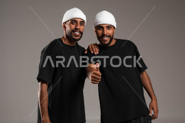إيماءات الاعجاب والموافقة، القبول و الرضا بالنتيجة، لاتفاق على أمر معين، النظر إلى الكاميرا بتعابير السرور، بورتريه مقرب لأخين توءم عربيين خليجيين سعوديين يرتديان الزي الكاجوال احدهما يرفع اصبع الابهام للأعلى بعلامة الجودة، خلفية رمادية