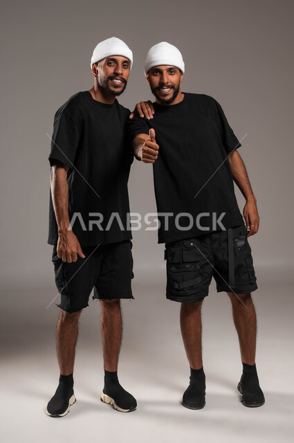 القبول و الرضا بالنتيجة، لاتفاق على أمر معين، النظر إلى الكاميرا بتعابير السرور، ايماءات الاعجاب والموافقة، بورتريه لأخين توءم عربيين خليجيين سعوديين يرتديان الزي الكاجوال احدهما يرفع اصبع الابهام للأعلى بعلامة الجودة، صورة لكامل الجسم، خلفية رمادية