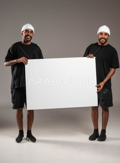 Two young Saudi men holding in their hand a blank white billboard looking at the camera with a smile, using illustrative panels, portrait of two men, smiling Saudi Gulf Arab twins wearing casual clothes holding a large rectangular mockup, full length body portrait, gray background