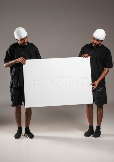 استخدام لوحات توضيحية و وسائل تعليمية، لنظر إلى الكاميرا بابتسامة، لوح اعلاني فارغ، إعلانات وعروض تسويقية، بورتريه لشابان توءم عربيين خليجيين سعوديين يرتديان الزي الكاجوال و قبعة بيضاء يحملان موك اب مستطيل كبير، صورة الجسم بالطول الكامل، خلفية رمادية