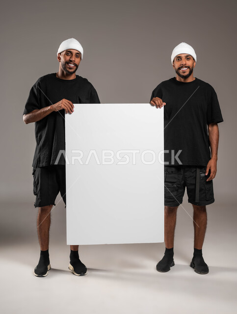 النظر إلى الكاميرا بابتسامة، لوح اعلاني فارغ، استخدام لوحات توضيحية ووسائل تعليمية، إعلانات وعروض تسويقية، بورتريه لشابان توءم عربيين خليجيين سعوديين يرتديان الزي الكاجوال و قبعة بيضاء يحملان موك اب مستطيل كبير، صورة الجسم بالطول الكامل، خلفية رمادية