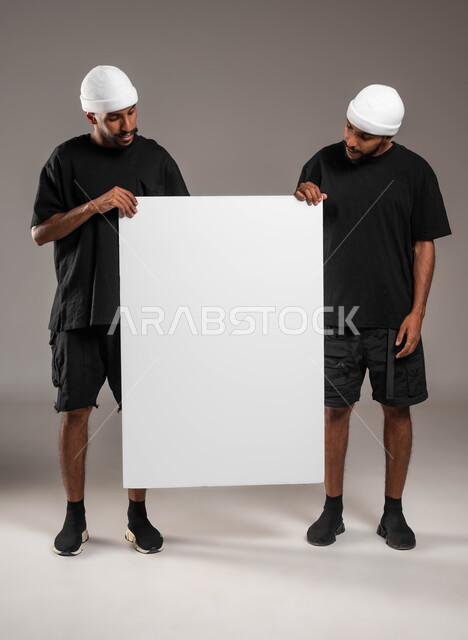 لوح اعلاني فارغ، إيماءات السرور والسعادة، استخدام لوحات توضيحية ووسائل تعليمية، إعلانات وعروض تسويقية، بورتريه لشابان توءم عربيين خليجيين سعوديين يرتديان الزي الكاجوال و قبعة بيضاء يحملان موك اب مستطيل كبير، صورة الجسم بالطول الكامل، خلفية رمادية