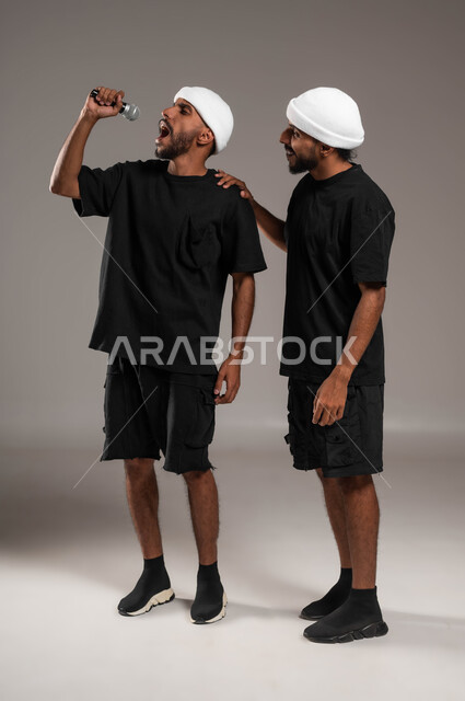 التفاعل و الاندماج مع الموسيقى، بورتريه لشابين توءم عربيين خليجيين سعوديين يرتديين الزي الكاجوال يقومان بعرض غنائي مسرحي، الاستمتاع بممارسة هواية الغناء باستخدام الميكرفون، تقديم المواهب الصوتية بإيماءات الحماس والسرور، صورة لكامل الجسم، خلفية رمادية