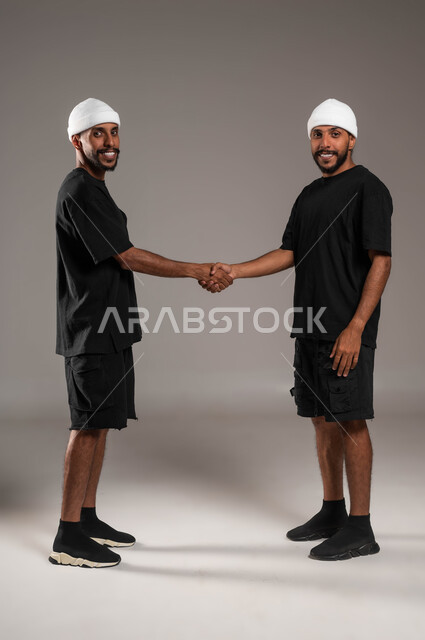 الاتفاق و الشراكة على المشاريع و الاعمال التجارية، مفهوم العلاقات الاجتماعية، لقاء العائلة والاصدقاء والتواصل، المحبة والترابط بين افراد الأسرة، التعارف واتفاقيات العمل، بورترية لشابين عربيين سعوديين خليجيين يرتديان الزي الكاجوال، صورة لكامل الجسم، خلفية رمادية