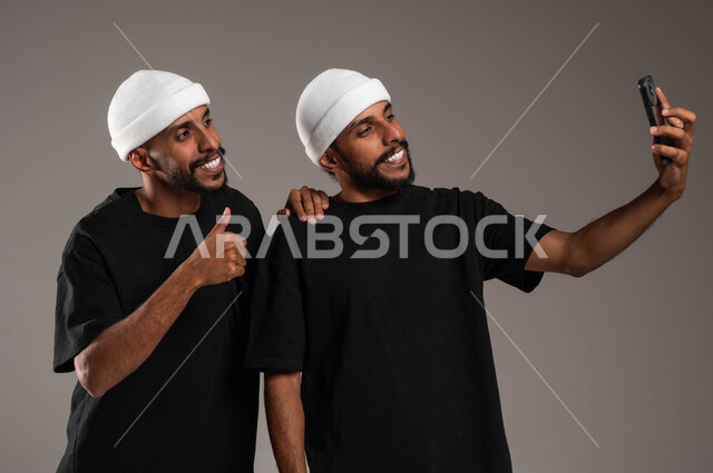 استخدام الجوال لالتقاط صور سيلفي تذكارية، بورتريه مقرب لشابين توءم عربيين سعوديين خليجيينن يرتديان الزي الكاجوال احدهما يمسك الهاتف المحمول بيده والآخر يرفع اصبعه بعلامة الجودة والامتياز، إيماءات السرور و السعادة، خلفية رمادية