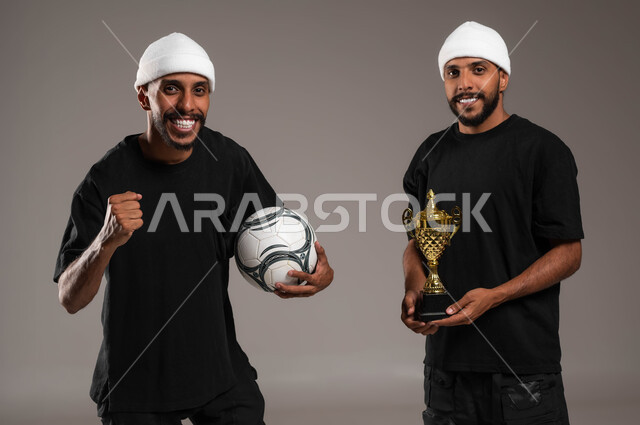 Entertaining sports activities, the concept of winning and being victorious in matches, gestures of joy and happiness, a feeling of joy and pleasure, a close-up portrait of two young Saudi Gulf Arab twins wearing casual clothes, one of them holding a football and the other holding a championship cup, gray background.