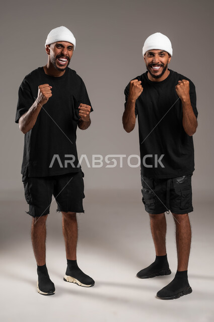 Raising hands high with gestures of victory, success and achieving goals, the concept of happiness in achieving goals, feelings of love and friendliness between brothers, a feeling of joy and happiness, a portrait of two young Saudi Gulf Arab twins wearing casual clothes and a white hat, a full-body portrait, gray background.