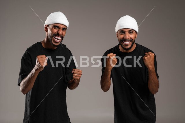 النظر للكاميرا بإيماءات الفوز والوصول للغايات، رفع اليدين عاليا بتعابير النجاح تحقيق الأهداف، مشاعر المحبة والود بين الاخوة، الشعور بالفرح والسرور، بورتريه مقرب لشابين توءم عربيين خليجيين سعوديين مبتسمين يرتديان الزي الكاجوال و قبعة بيضاء، خلفية رمادية
