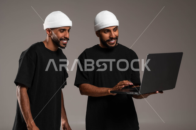 استخدام الأجهزة الالكترونية الحديثة,  استخدام الحاسوب في تطوير العمل, دور التقنيات المتطورة في ايجاد المعلومات, بورتريه مقرب لشابان عربيان سعوديان خليجيان يرتديان الزي الكاجول و يضعان قبعة بيضاء احدهما يمسك بيده الابتوب, خلفية رمادية