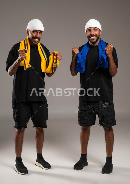 رفع اليدين بإيماءات الفوز ، النظر الى الكاميرا بإيماءات الحماس و الشغف، المباريات والألعاب الرياضية، تعابير الفرح  والسرور لانتصار الفريق، بورتريه لشابين توءم عربيين خليجيين سعوديين يرتديين أوشحة تشجيعية لفرق كرة القدم، صورة لكامل الجسم، خلفية رمادية