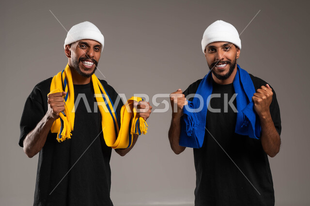 النظر الى الكاميرا بإيماءات الحماس و الشغف، المباريات والألعاب الرياضية، تعابير الفرح  والسرور لانتصار الفريق، بورتريه مقرب لشابان توءم عربيان خليجيان سعوديان يرتديان أوشحة تشجيعية لفرق كرة القدم، رفع اليدين بتعابير الفوز، خلفية رمادية