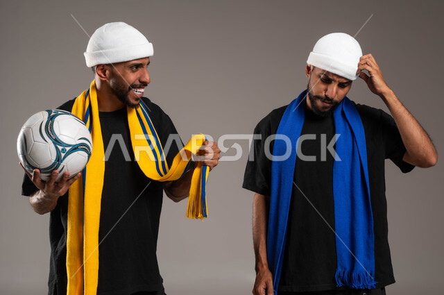 تعابير الفرح بالنصر والفوز، إيماءات الحزن لخسارة الفريق، بورتريه مقرب لشاب عربي خليجي سعودي يقوم بإغاظة أخوه التوأم لانتصار فريقه المفضل يحمل بيده كرة قدم، ارتداء اوشحة تشجيعية لفرق كرة القدم، المباريات والألعاب الرياضية، خلفية رمادية