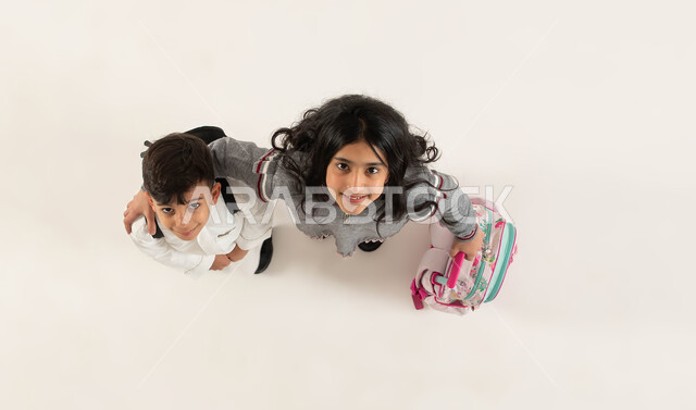The concept of a friendly relationship between sisters, preparing for the new school year with gestures indicating happiness, a vertical overhead portrait of two Saudi Gulf Arab brothers carrying a school bag looking at the camera, education in Saudi Arabia and perseverance in studying to achieve success, white background