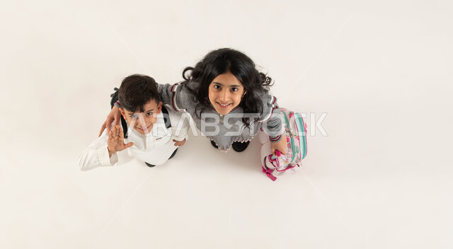 العلاقة الودية بين الأخوات, الاستعداد للعام الدراسي الجديد بايماءات تدل على السعادة، بورتريه علوي عامودي لأخوين عربيين سعوديين خليجيين يحملان الحقيبة المدرسية ينظران الى الكاميرا, مفهوم التعليم في السعودية والمثابرة على الدراسة لتحقيق النجاح, خلفية بيضاء