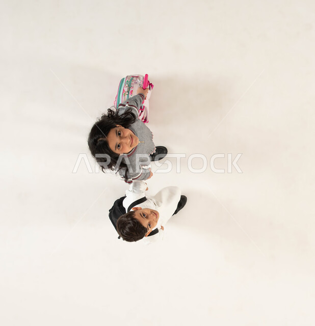 The concept of education in Saudi Arabia and perseverance in studying to achieve success, preparing for the new academic year with gestures indicating joy and happiness, a vertical portrait of a Saudi Gulf Arab boy and girl carrying a school backpack, going to school, white background