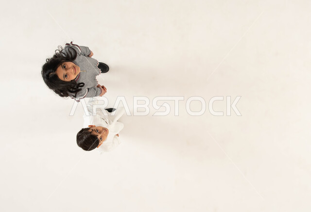 تقوية العلاقة الإيجابية بين الأخوة, بورتريه علوي عامودي لأخويين عربيين سعوديين خليجيين يمسكان بيد بعضهما, مفهوم الألفة والمحبة بين الأشقاء, الاستمتاع بقضاء أوقات ممتعه برفقة الأخوات, النظر الى الكاميرا بايماءات السعادة والسرور, خلفية بيضاء