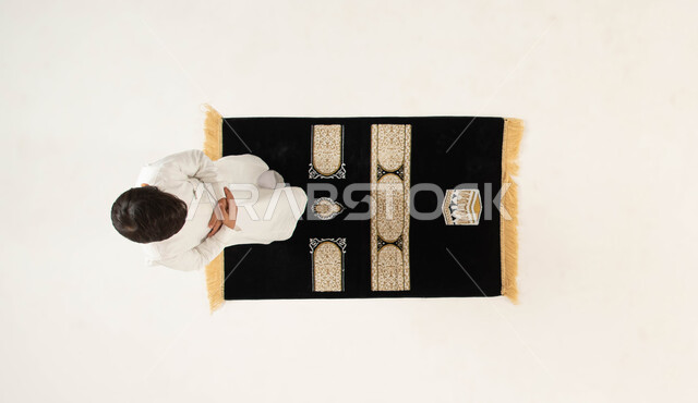 The importance of teaching children to commit to praying on time, getting closer to God Almighty by performing the obligations and adhering to His commands, a vertical overhead portrait of a Saudi Gulf Arab boy wearing the traditional dress and praying to God Almighty, the concept of the pillars of Islam, Islamic matters and worship, white background