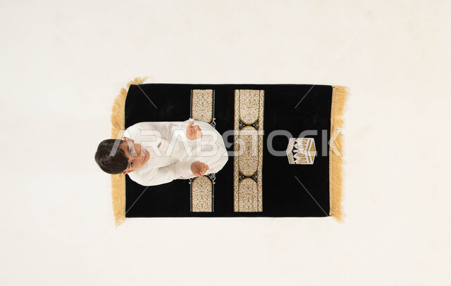 الجلوس على سجادة الصلاة والقيام بالأذكار والأدعية اليومية, الدعاء والتقرب من الله تعالى, بورتريه علوي عامودي لطفل عربي خليجي سعودي مبتسم يرتدي الثوب التقليدي يقوم بالدعاء الى الله ينظر الى الكاميرا, مفهوم أركان الإسلام, إسلاميات وعبادة, خلفية بيضاء