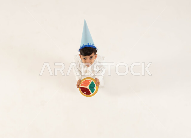 الاستمتاع بالمناسبات وأعياد الميلاد, بورتريه علوي عامودي لولد عربي سعودي خليجي مبتسم يرتدي قبعة الإحتفال بعيد الميلاد ويحمل قالب الكيك المزين بالشموع ينظرالى الكاميرا, ايماءات السعادة والسرور بقضاء اوقات ممتعة, خلفية بيضاء