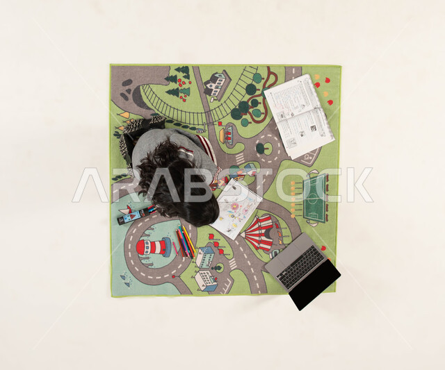 Integrating the educational process with technique and technology, developing artistic talents, learning and receiving lessons remotely, a vertical overhead portrait of a Saudi Arabian Gulf girl sitting on carpets with a modern design, drawing, coloring, and doing homework, white background.