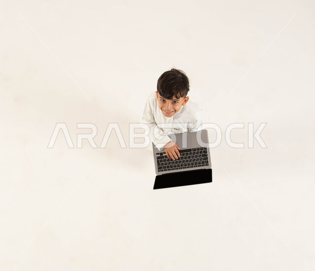 دورات تعلمية ثقافية و انشطة ترفيهية الكترونية للاطفال، التعلم وتلقي دروس الاونلاين عن بعد، بورتريه علوي عامودي لولد عربي خليجي سعودي مبتسم يرتدي الزي التقليدي يحمل جهاز الحاسوب ينظر الى الكاميرا، قضاء اوقات مسلية باللعب في الحاسب المحمول، خلفيه بيضاء