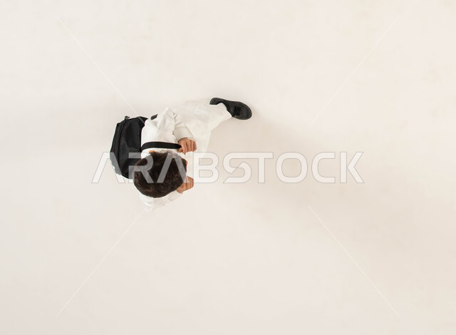 التحضير لبداية عام دراسي جديد، الاستعداد والتجهز للذهاب الى المدرسة، بورتريه علوي عامودي لطالب عربي خليجي سعودي مبتسم يرتدي الزي التقليدي يحمل حقيبه الظهر المدرسيه، ايماءات السعادة و السرور للعودة الى الدراسة، خلفيه بيضاء .