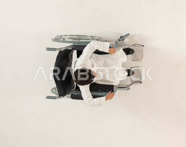 الضعف في القدرة الحركية، تحسين حياة أصحاب الهمم وتأمين احتياجاتهم الخاصة، شعور المعاقين بالإنتماء والتقدير، بورتريه علوي عامودي لولد عربي خليجي سعودي يرتدي الزي التقليدي من ذوي الاحتياجات الخاصه يجلس على كرسي متحرك، خلفيه بيضاء