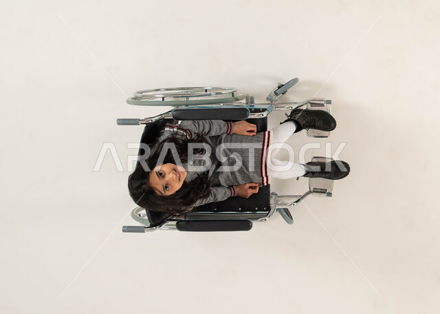 الضعف في القدرة الحركية، تحسين حياة أصحاب الهمم وتأمين احتياجاتهم الخاصة، شعور المعاقين بالإنتماء والتقدير والإحترام في السعوديه، بورتريه علوي عامودي لفتاة عربيه خليجيه سعودية من ذوي الاحتياجات الخاصه تجلس على كرسي متحرك و تنظر الى الكاميرا، خلفيه بيضاء