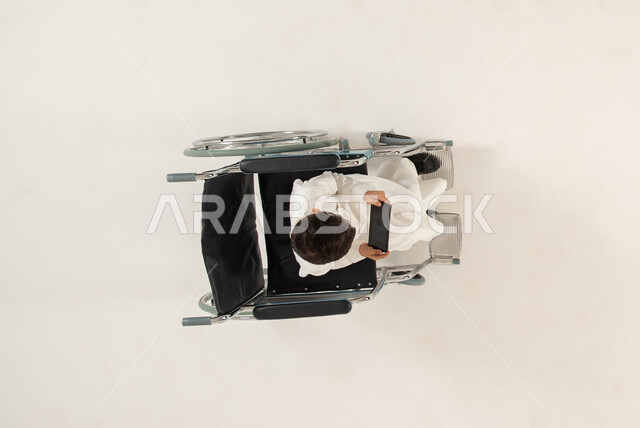 عرض شاشه سوداء فارغه عبر الجوال، العنايه بأصحاب الهمم وتأمين احتياجاتهم الخاصة، قضاء اوقات ممتعة ومسلية، بورتريه علوي عامودي لولد عربي خليجي سعودي مقعد من ذوي الاحتياجات الخاصه يجلس على كرسي متحرك ويحمل بيده الهاتف المحمول، خلفيه بيضاء .