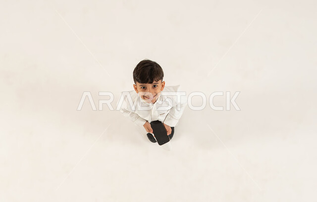 استخدام التقنيات الحديثة المتطورة، عرض شاشة سوداء فارغه عبر الهاتف المحمول، بورتريه علوي عامودي لولد عربي خليجي سعودي مبتسم يرتدي الزي التقليدي يحمل بيده الجوال وينظر الى الكاميرا بإيماءات السرور، خلفيه بيضاء .