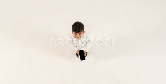 عرض شاشة سوداء فارغه عبر الهاتف المحمول، بورتريه علوي عامودي لولد عربي خليجي سعودي مبتسم يرتدي الزي التقليدي يحمل بيده الجوال وينظر الى الكاميرا بإيماءات السرور، استخدام التقنيات الحديثة المتطورة، خلفيه بيضاء .