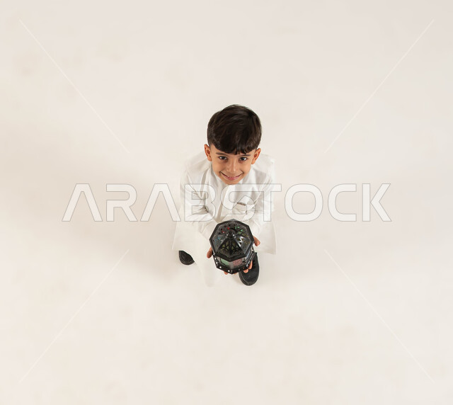 Religious and Islamic holidays and occasions, decoration and preparation for the spiritual atmosphere of Ramadan, preparations to welcome the holy month of Ramadan, a vertical overhead portrait of a smiling Saudi Gulf Arab boy holding a metal lantern in his hand and looking at the camera, white background.