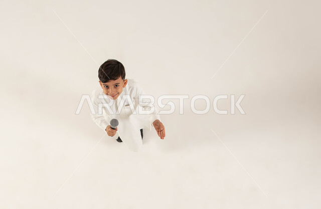 Discovering singing talents in children from a young age, a vertical portrait of a smiling Saudi Arabian Gulf boy holding a microphone in his hand and looking at the camera, presenting theatrical and singing artistic performances and entertaining entertainment segments, gestures of enthusiasm to begin vocal training, a white background.