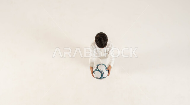 ممارسة الهوايات المفضلة ،تطوير المهارات البدنية ،بورتريه علوي عامودي لولد عربي خليجي سعودي يرتدي الزي التقليدي يمد يديه الى الأمام ويحمل الكرة، انشطة رياضية ترفيهيه للأطفال، الشغف في متابعه فريق كرة القدم منذ الصغر، خلفيه بيضاء .