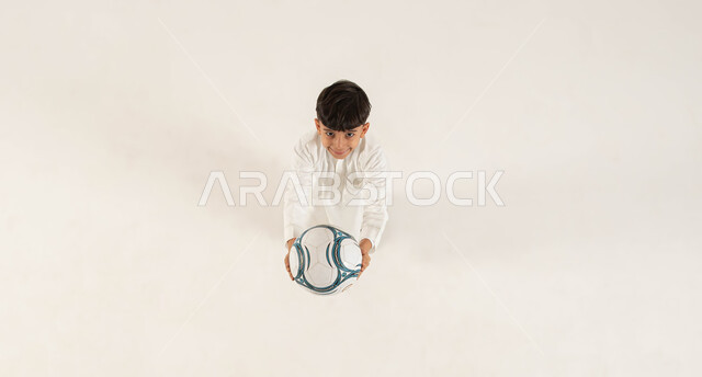 النظر الى الكاميرا بايماءات السرور، تعابير الحماس للفوز في المباراة، بورتريه علوي عامودي لولد عربي خليجي سعودي مبتسم يرتدي الزي التقليدي يمد يديه للأمام و يحمل الكرة ،انشطة رياضية ترفيهيه للأطفال، الشغف في متابعه فريق كرة القدم منذ الصغر، خلفيه بيضاء .