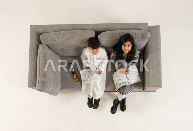 Developing artistic and mental skills, using free time with educational activities, a vertical portrait of a Saudi Gulf Arab boy and girl sitting on the sofa and looking at the camera with happy gestures, reading and reading useful books, spending an enjoyable time drawing and coloring, white background.