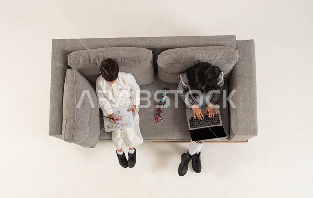 Spending an enjoyable time learning drawing and coloring, taking online lessons remotely via a laptop, a vertical overhead portrait of two Saudi Gulf Arab brothers sitting on the sofa taking advantage of their free time with useful electronic and real-life activities, a white background.
