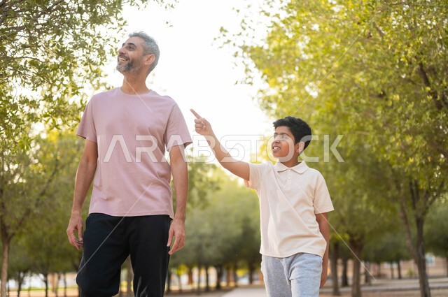 قضاء أوقات الإجازة مع الابناء، أجواء أسرية ممتعة، الترابط الأسري و الاستمتاع بالعطلة مع العائلة، مفهوم الرابطة الأبوية، الأشارة لمكان ما بإيماءات السعادة و السرور، أب عربي سعودي خليجي يرتدي الزي الكاجوال يتنزه في الهواء الطلق برفقة ولده، خلفية الطبيعة