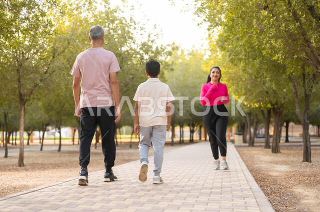 المشي في الحديقة و تبادل الأحاديث، قضاء لحظات ممتعة مع العائلة، الاهتمام بالصحة و المحافظة على اللياقة، أب عربي خليجي سعودي يرتدي الزي الكاجوال يتنزه في الهواء الطلق مع ابنه، امرأة عربية خليجية سعودية ترتدي ملابس رياضية تمارس رياضة الركض،  خلفية الطبيعة