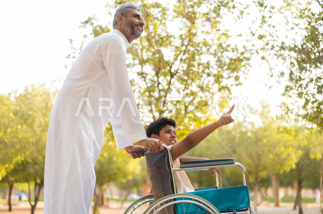 الجلوس على كرسي متحرك و الاشارة لشيء ما، قضاء الوقت في المنتزه مع العائلة، إيماءات المعاملة الحسنة، صورة مقربة من الجانب لأب عربي سعودي خليجي يرتدي الزي التقليدي يتنزه مع ابنه المقعد، دعم المجتمع لذوي الاحتياجات الخاصة، تأمين احتياجات أصحاب الهمم، خلفية الطبيعة